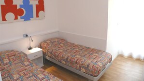 Een kamer met twee eenpersoonsbedden, een klein nachtkastje met een lamp, een aan de muur hangend kunstwerk en een raam met gordijnen.