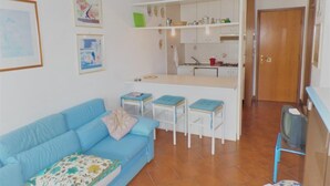 Una sala de estar con un sofá azul, una pequeña zona de cocina con armarios blancos y una puerta de madera.