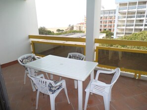 Un balcón con una mesa blanca y cuatro sillas blancas, una barandilla amarilla, y vistas a edificios y follaje.