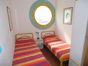 Una habitación pequeña con dos camas individuales, un ventanal redondo, una mesita de noche con lámpara y un cuadro enmarcado en la pared.