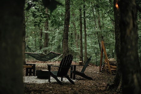 A hammock, a chair, and a small table in a forest setting.. Boho Farmhouse Camper with AC in Harpers Ferry