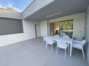 A modern outdoor patio with a white table and chairs, a sliding glass door leading to a balcony, and a view of greenery. - Bright beachside apartment with two terraces (Bibione)