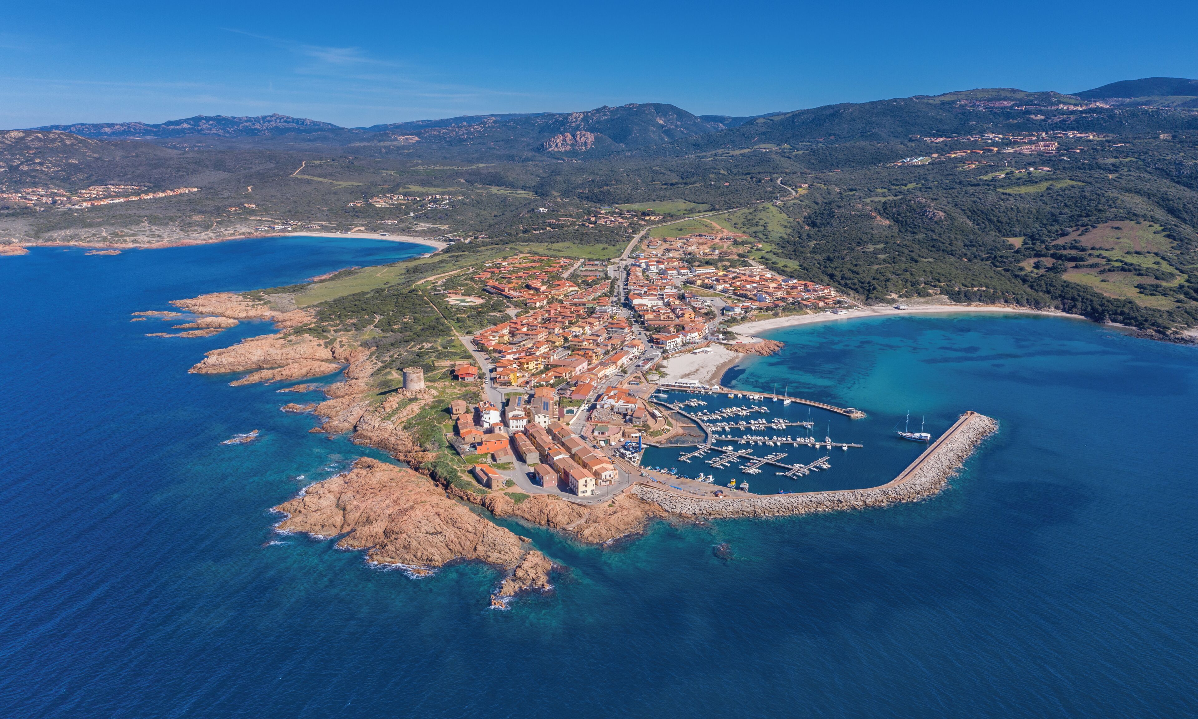 An aerial view of a coastal town with a marina, rocky terrain, and surrounding hills.