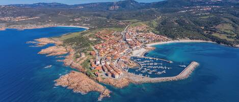 An aerial view of a coastal town with a marina, rocky terrain, and surrounding hills.