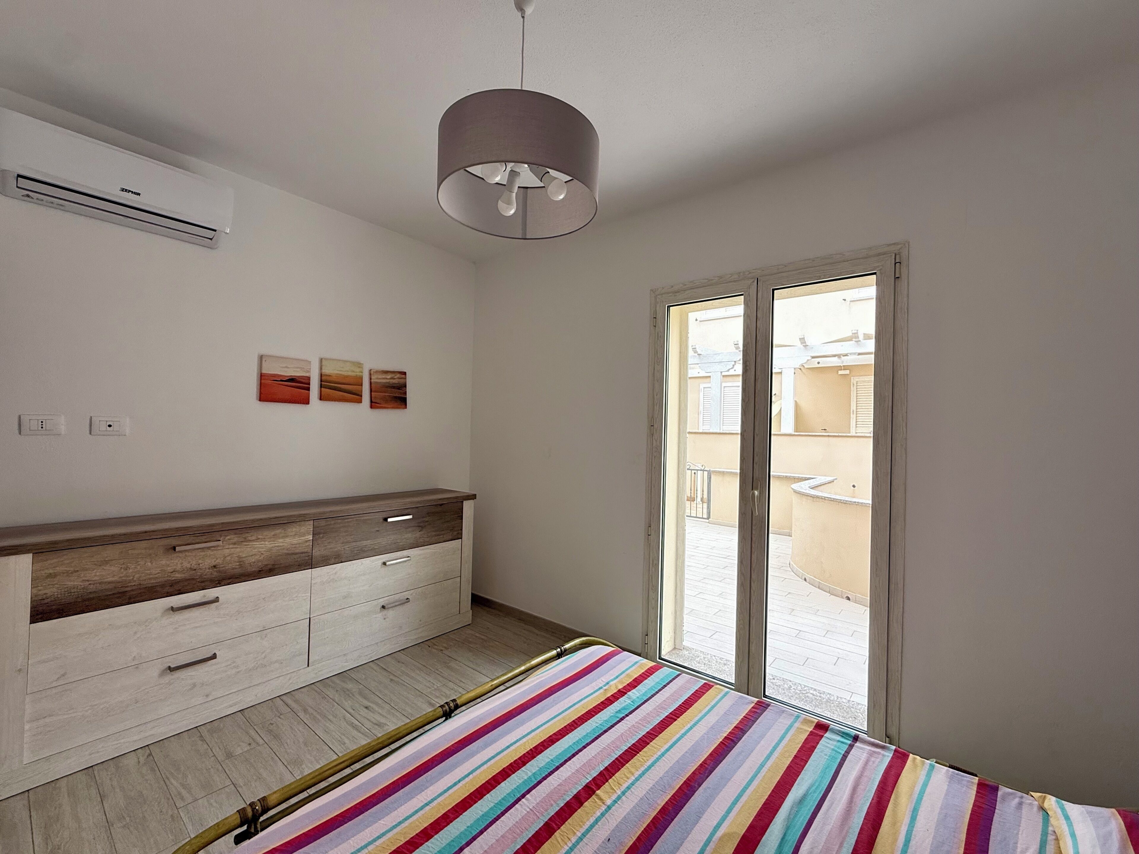 Un dormitorio con cama, mesita de noche, armario, luz de techo, ventana y vistas al exterior.