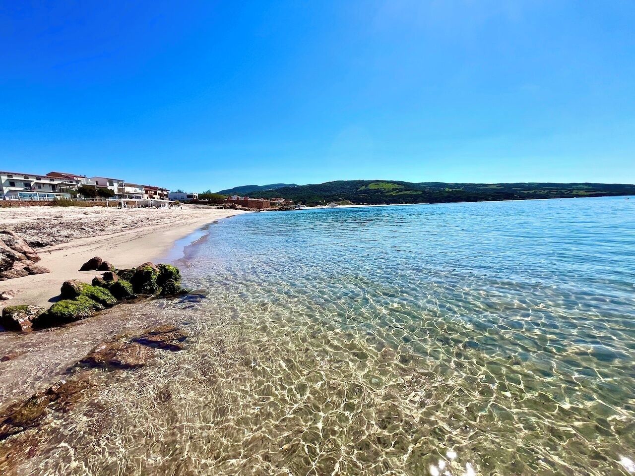 A beach with clear water, a rocky shoreline, and buildings in the background.