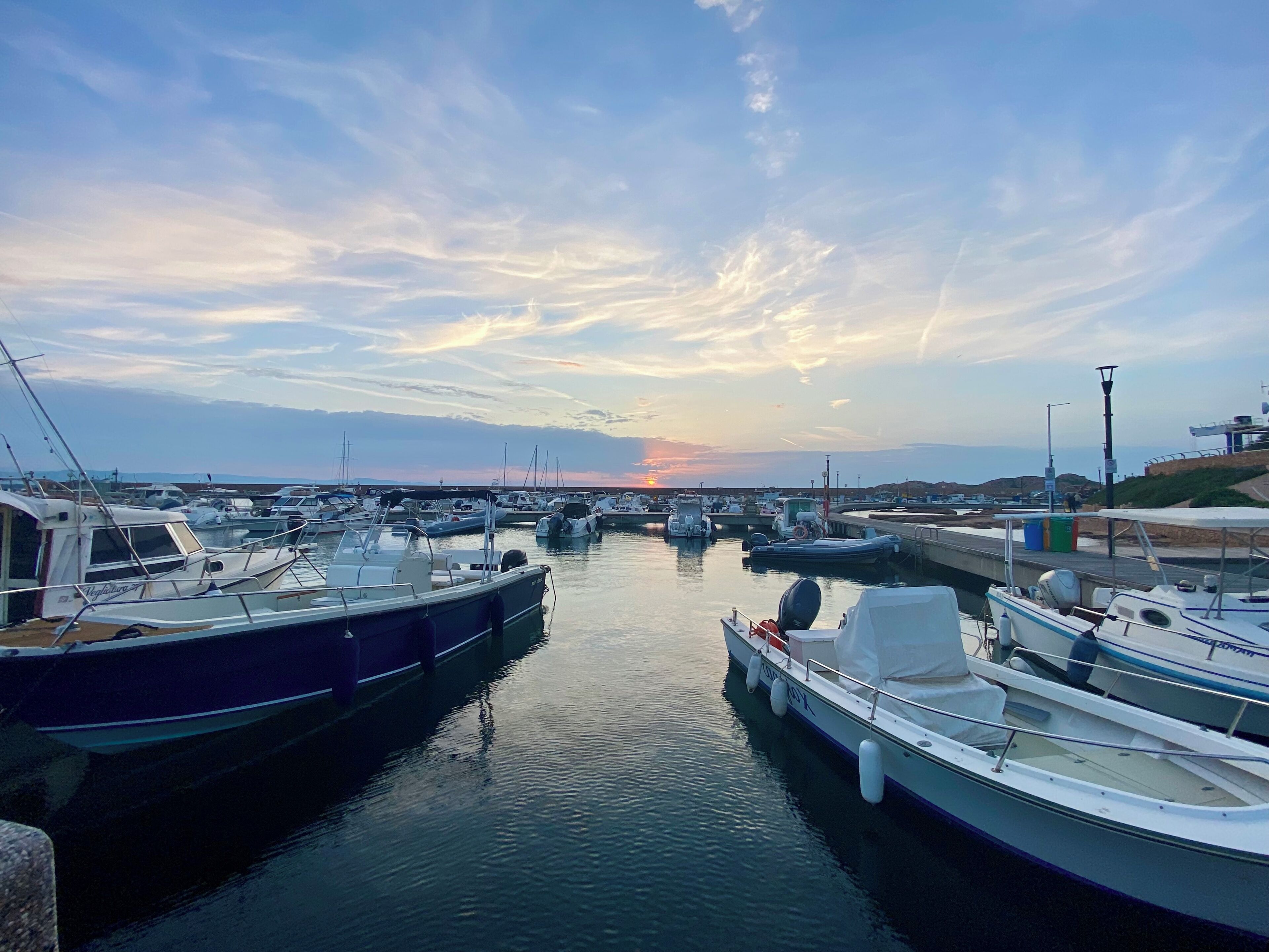 Un puerto deportivo con varias embarcaciones amarradas, agua tranquila y un atardecer al fondo.