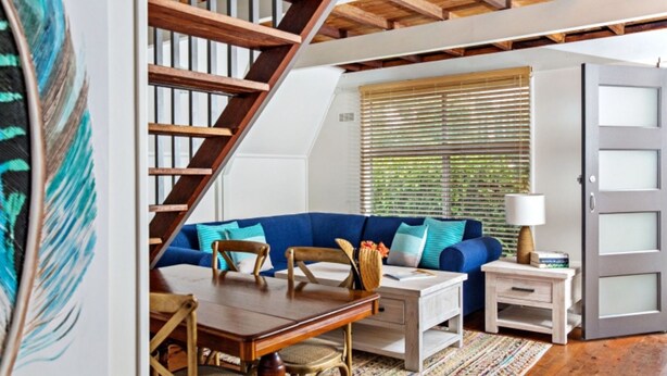 A room with a wooden staircase, a blue sofa, a wooden table, and a window with blinds. - Beach Chalet - Unique Triangular Beach House (Mollymook Beach)