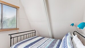 A bedroom with a sloped ceiling, a window with blinds, a bed with patterned bedding, and a desk lamp. - Beach Chalet - Unique Triangular Beach House (Mollymook Beach)