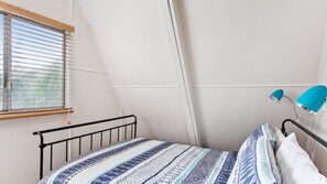 A bedroom with a sloped ceiling, a window with blinds, a bed with patterned bedding, and a desk lamp. - Beach Chalet - Unique Triangular Beach House (Mollymook Beach)