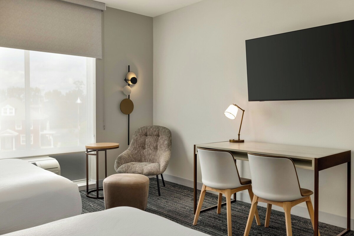 Habitación de hotel con cama, escritorio, dos sillas, una mesita redonda, lámpara y aplique de pared.