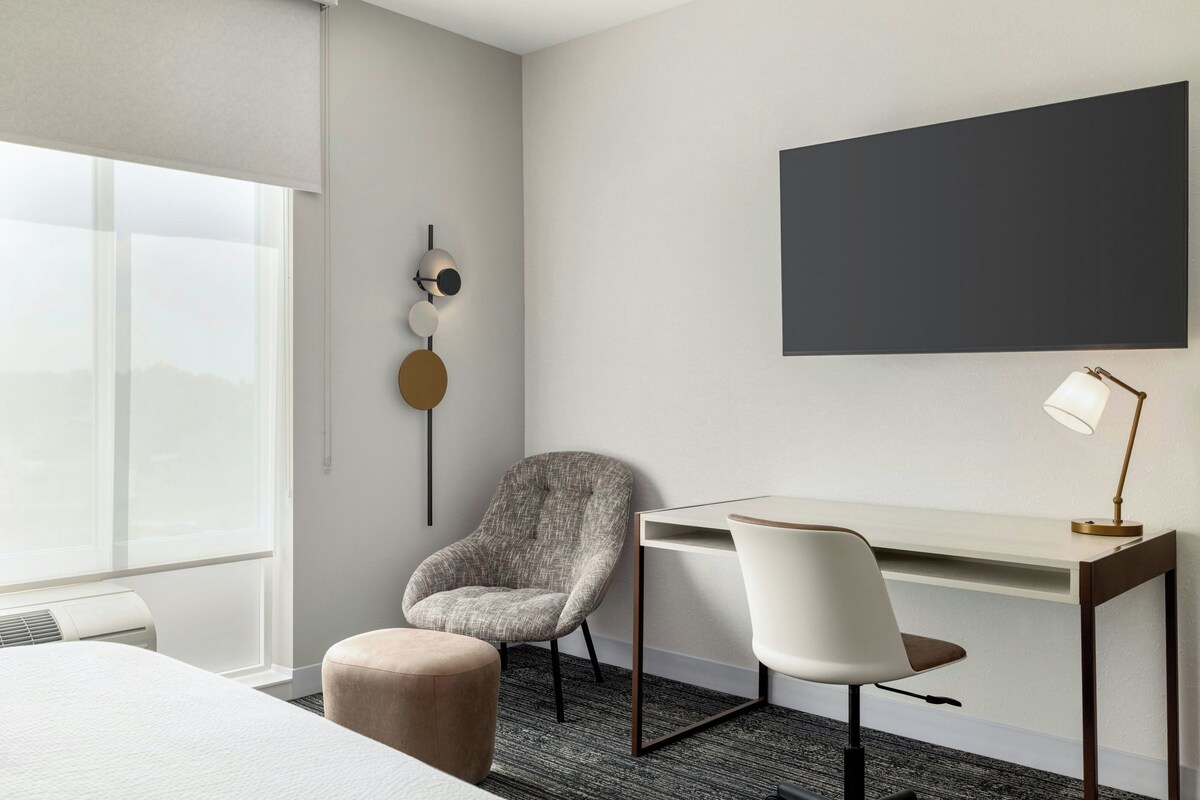 Una habitación de hotel con cama, escritorio, dos sillas, lámpara de pared y un ventanal grande.