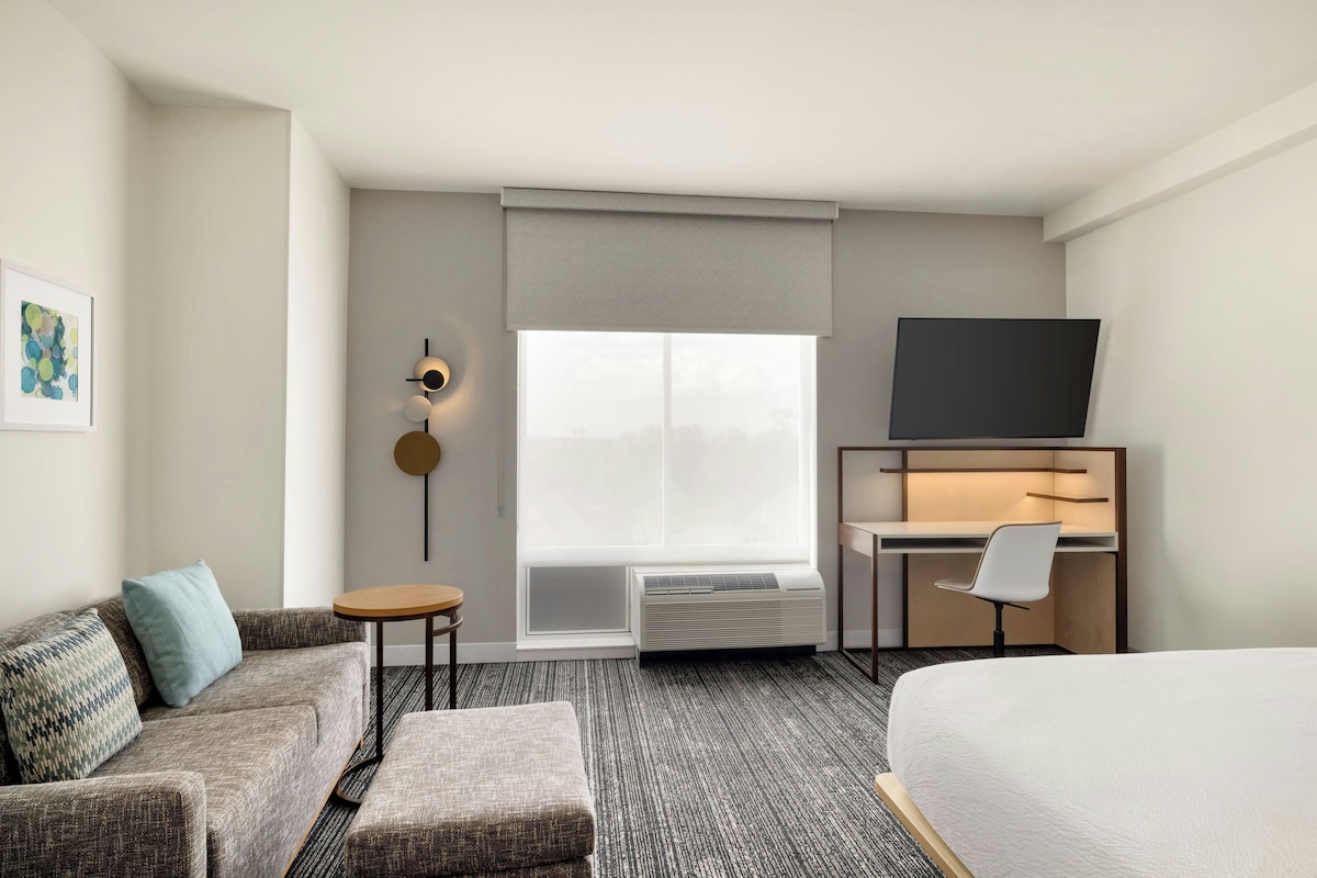 Una habitación de hotel con cama, sofá, escritorio con silla, televisor y ventana con persianas.