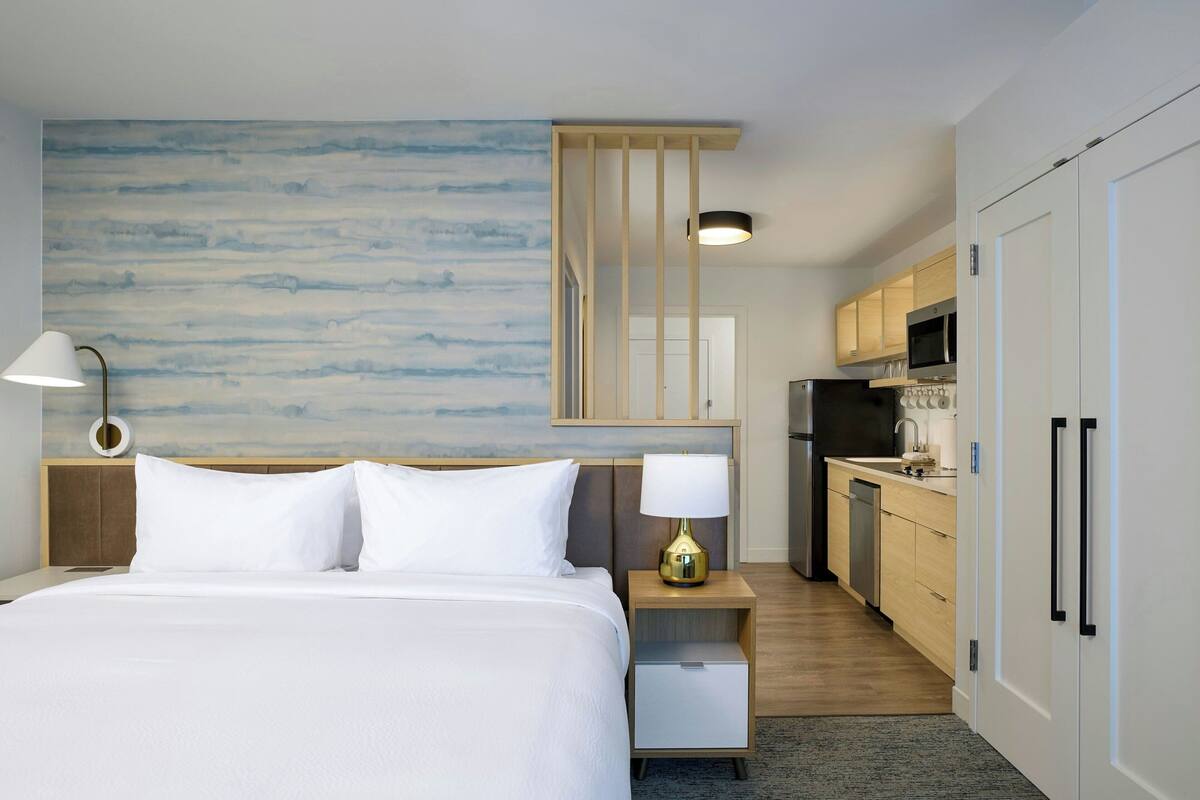 Habitación de hotel con cama, lámpara de noche, mesita de noche y vista al área de la cocina.