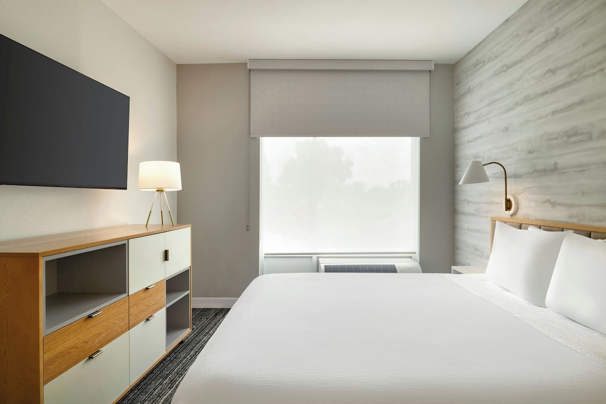 Una habitación de hotel con una cama, un escritorio con lámpara, un armario, un ventanal grande y una pared con textura similar al mármol.