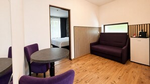A compact hotel room with a round table, purple chairs, a sofa, a bed, and a minibar.