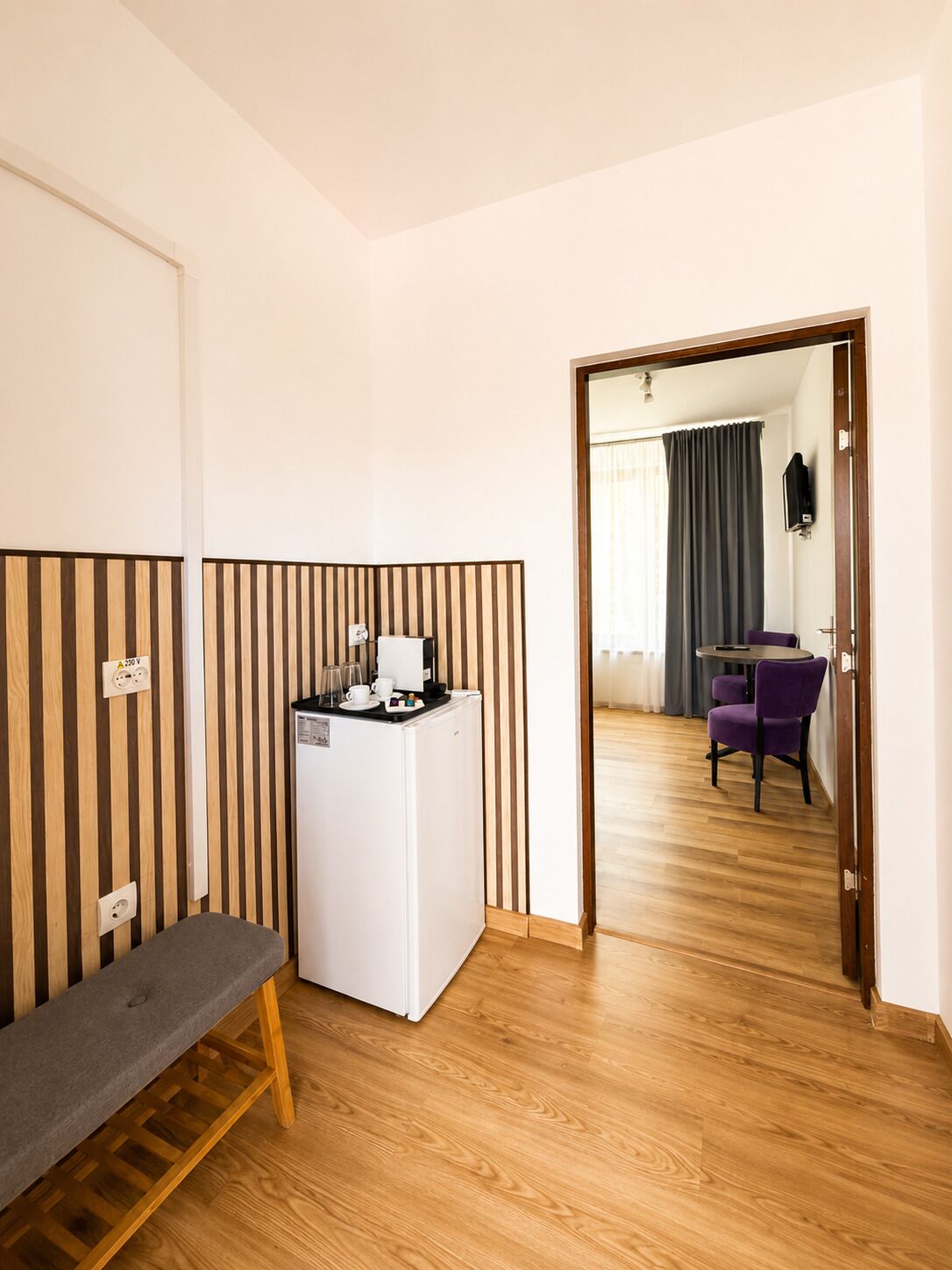Una habitación compacta con kitchenette, una banca, una mesa pequeña y una puerta que da a otra habitación con una silla morada.