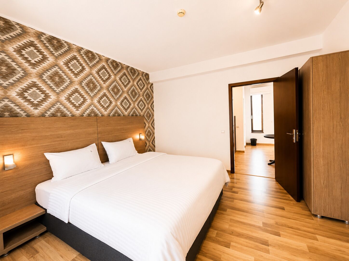 Une chambre d’hôtel avec un grand lit, une tête de lit en bois, un papier peint à motifs et une vue sur le couloir.