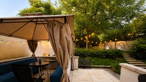 An outdoor patio with a blue sofa, a table, and chairs. There is a pergola with a curtain, string lights, and a staircase leading to a higher level.