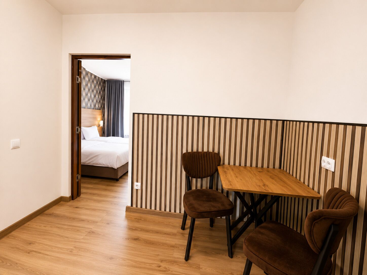 A hotel room with a wooden floor, a bed with white linens, a striped wall panel, and a small dining table with two chairs.