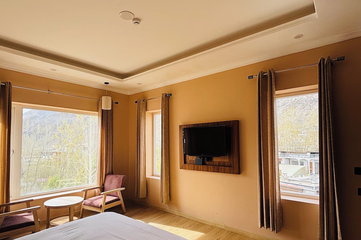 Een hotelkamer met een bed, een aan de muur bevestigde televisie, een klein rond tafeltje, een stoel en grote ramen met gordijnen.