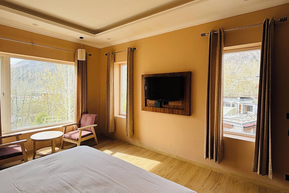 Hotelkamer met een bed, een aan de muur bevestigde televisie, een stoel, een kleine tafel en grote ramen met gordijnen.