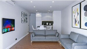 A modern living room with a grey sofa, a flat-screen TV, and a kitchen area in the background. - Budget VictoryS Ensuite Room (Gibraltar)