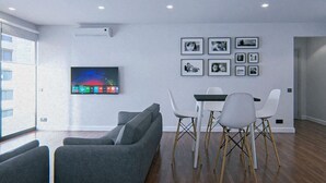 A modern living room with a flat-screen TV, grey sofa, white dining chairs, and framed pictures on the wall. - Budget VictoryS Ensuite Room (Gibraltar)
