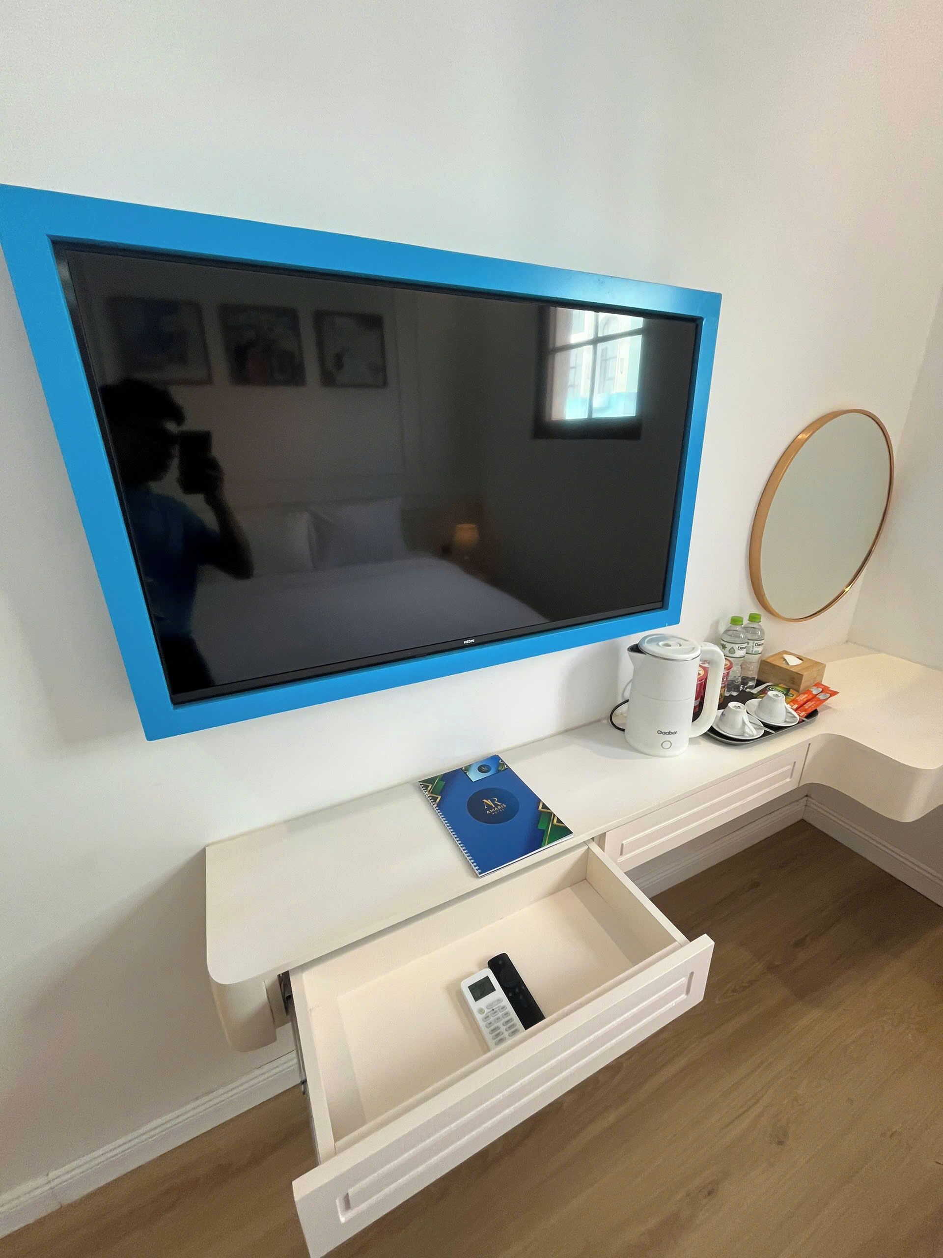 A hotel room with a large flat-screen TV, a bedside table with a coffee maker, a mirror, and a drawer with a remote control.