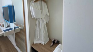 Habitación de hotel con piso de madera, una bata blanca colgada en un perchero, un espejo, un escritorio con computadora y un refrigerador negro.