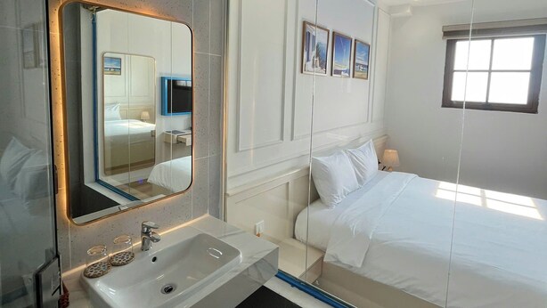 Una habitación de hotel moderna con un espejo grande, una cama blanca, una mesita de noche, una ventana y un baño con lavamanos y espejo.