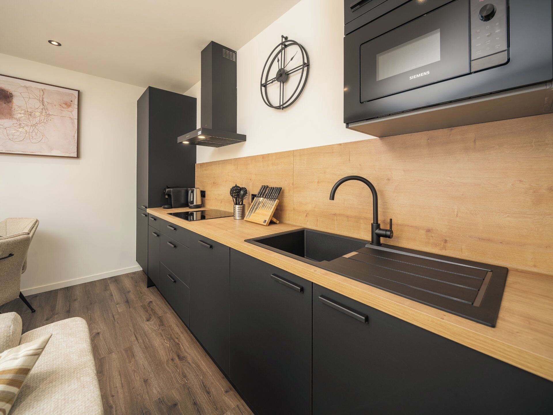 Une cuisine moderne avec des meubles de couleur noire, un plan de travail en bois, un évier, un four à micro-ondes et une horloge murale.