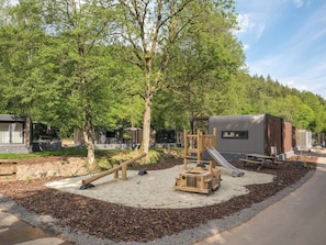 A playground with a slide and climbing structures, surrounded by trees and a gravel path.