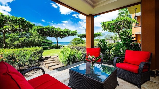 An outdoor seating area with wicker furniture and red cushions, overlooking a lush garden and the ocean. - Ground floor Condo Steps from the Sand with Pool and Sunset Views (Playa Langosta)