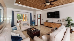 A modern living room with a ceiling fan, a flat-screen TV, and a large potted plant. - Ground floor Condo Steps from the Sand with Pool and Sunset Views (Playa Langosta)