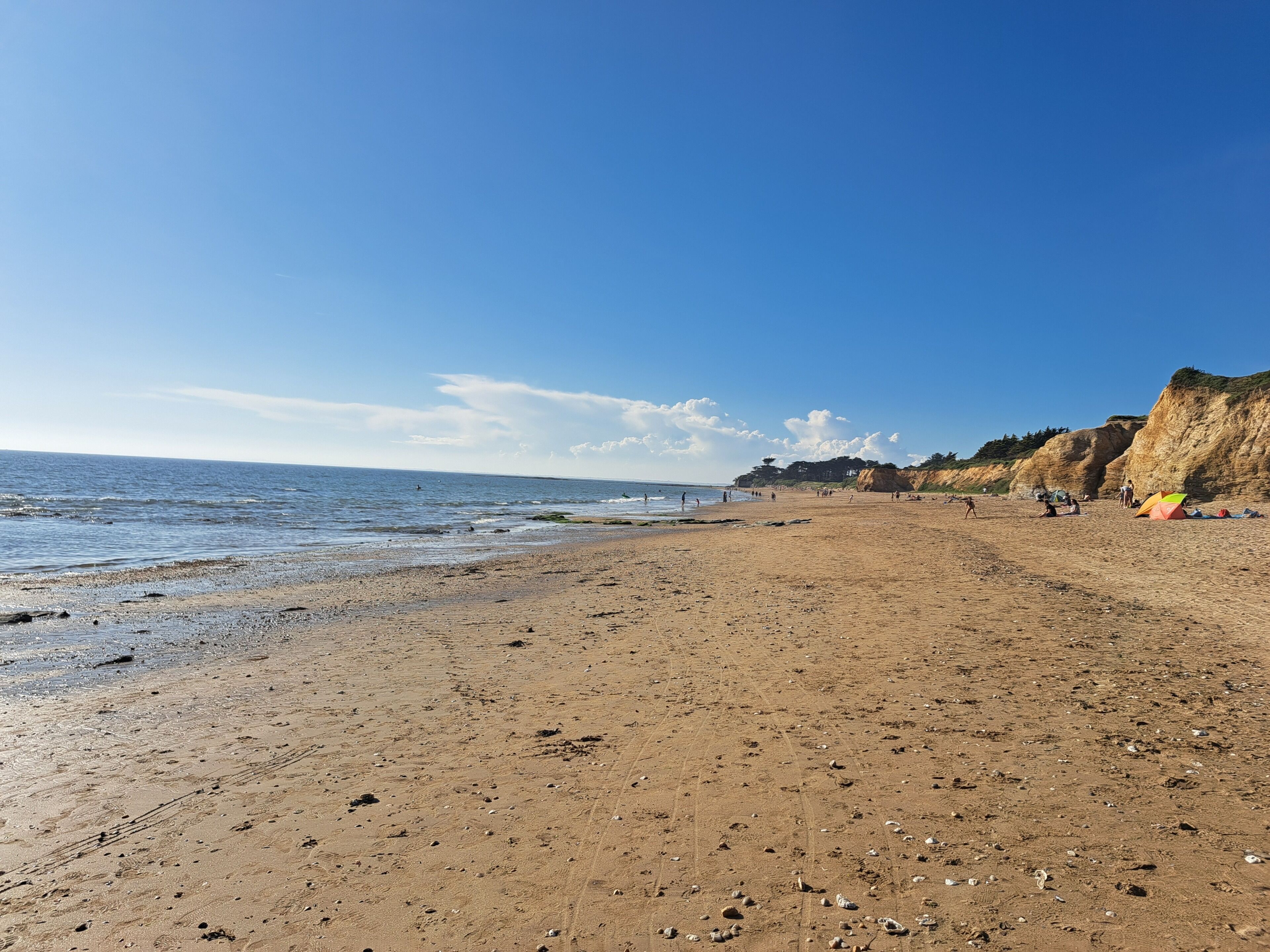 Ein weiter Sandstrand mit blauem Himmel, felsigen Klippen und einigen Personen sowie Zelten in der Ferne.