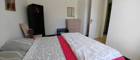 A bedroom with a bed, a wardrobe, a dresser, a mirror, and a door leading to another room.
