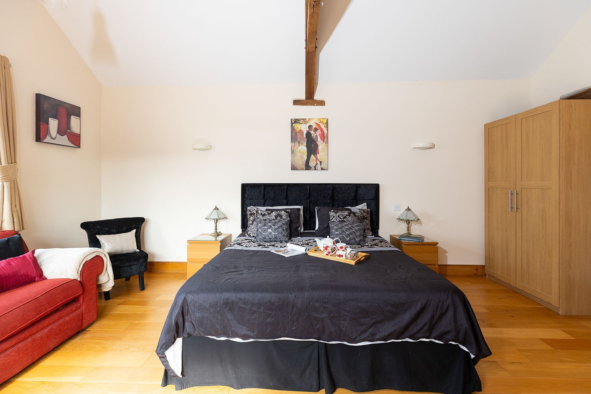 Un dormitorio con una cama grande, dos mesitas de noche, un sofá rojo, un armario de madera y obras de arte en las paredes.