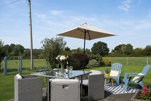 Eine Terrasse im Freien mit einem Glastisch, Korbstühlen, einem beigen Sonnenschirm und einer Vase mit Blumen.