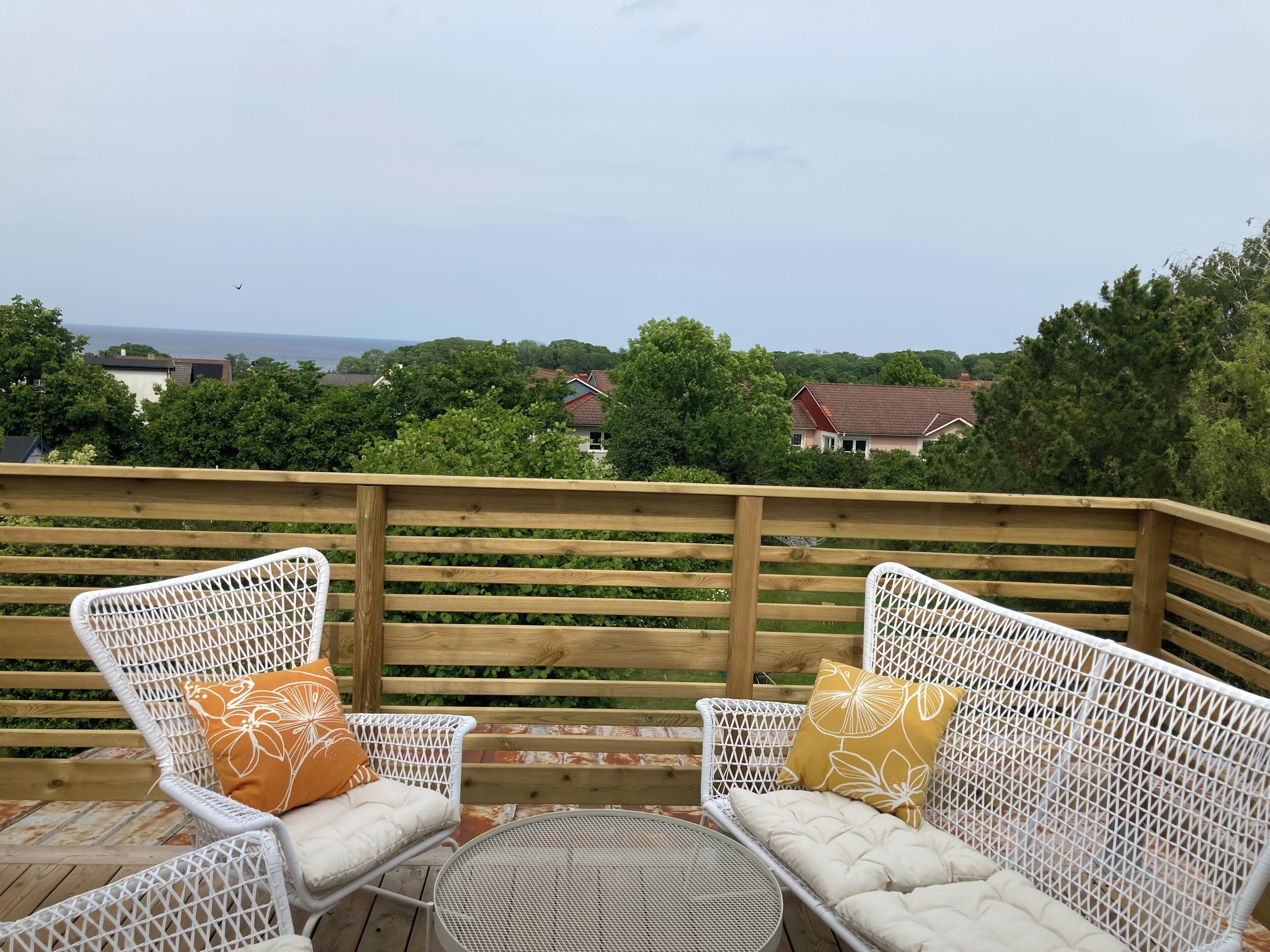 Een houten terras met rieten stoelen en een ronde tafel, met uitzicht op een woonwijk met huizen en bomen.