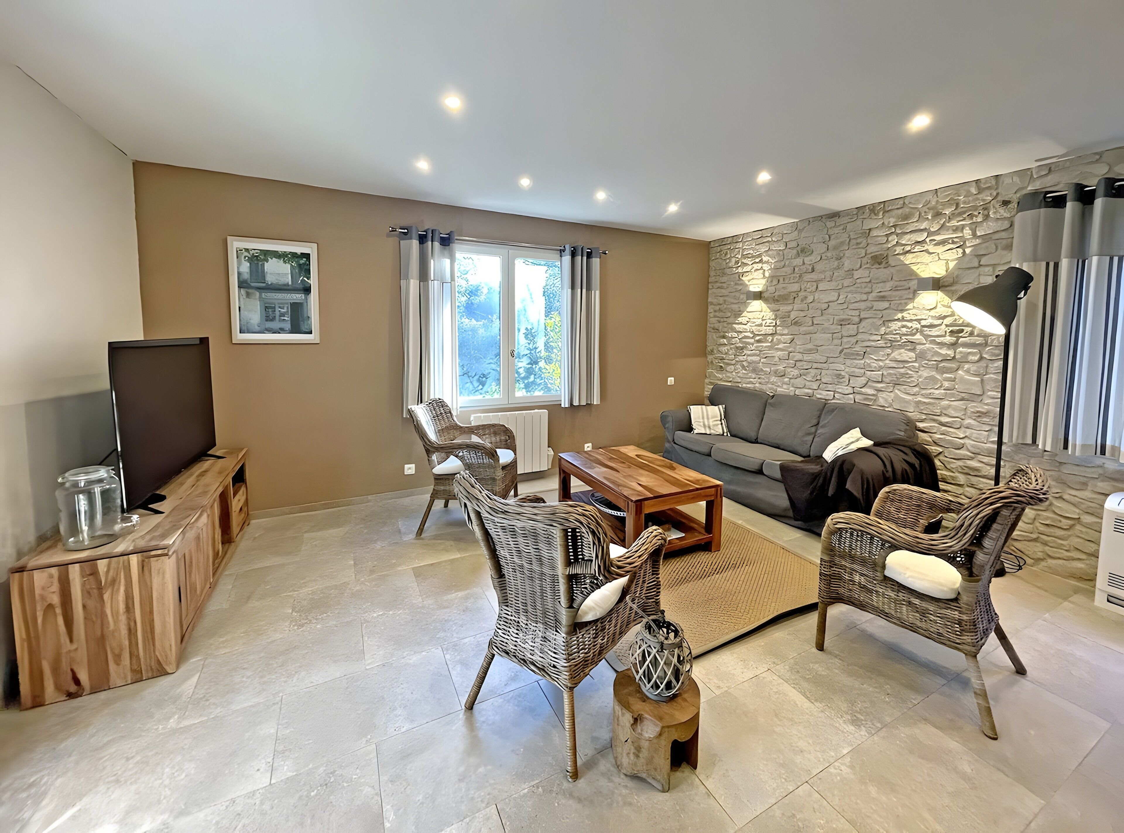Een woonkamer met een bank, twee rieten fauteuils, een houten salontafel, een televisie op een houten dressoir, een stenen muur en een raam met gordijnen.