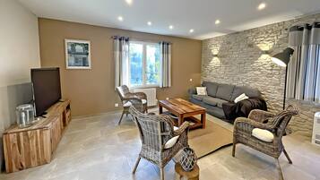 Een woonkamer met een bank, twee rieten fauteuils, een houten salontafel, een televisie op een houten dressoir, een stenen muur en een raam met gordijnen.
