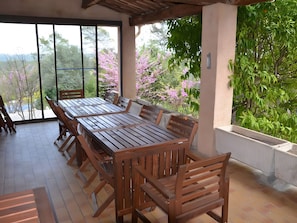 Un espace de restauration extérieur abrité, doté d’une grande table et de chaises en bois, entouré de grandes fenêtres et d’une végétation luxuriante.