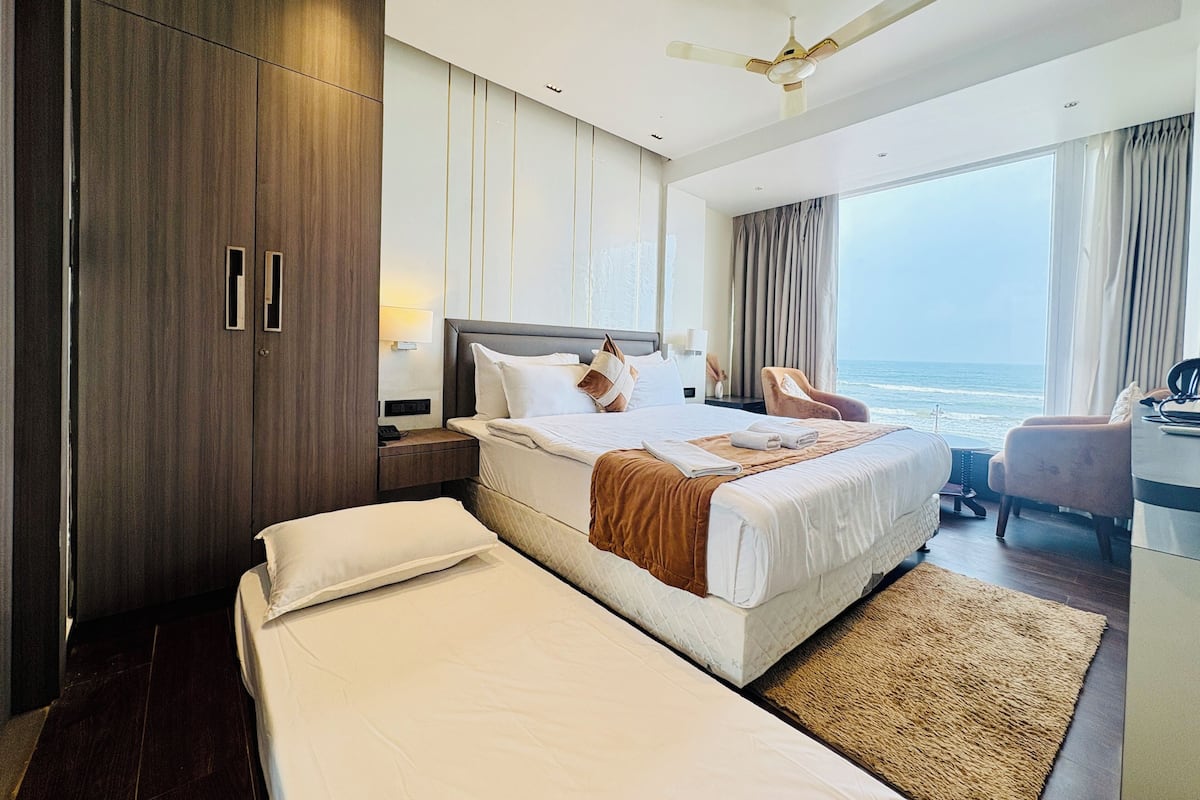 A modern hotel room with a large bed, a separate sofa, a desk, and a view of the ocean.