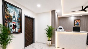 A modern hotel lobby with a reception desk, a painting of a couple walking, a potted plant, and a ceiling fan. - Mauryan Signature Hotel NHPC Metro (Faridabad)
