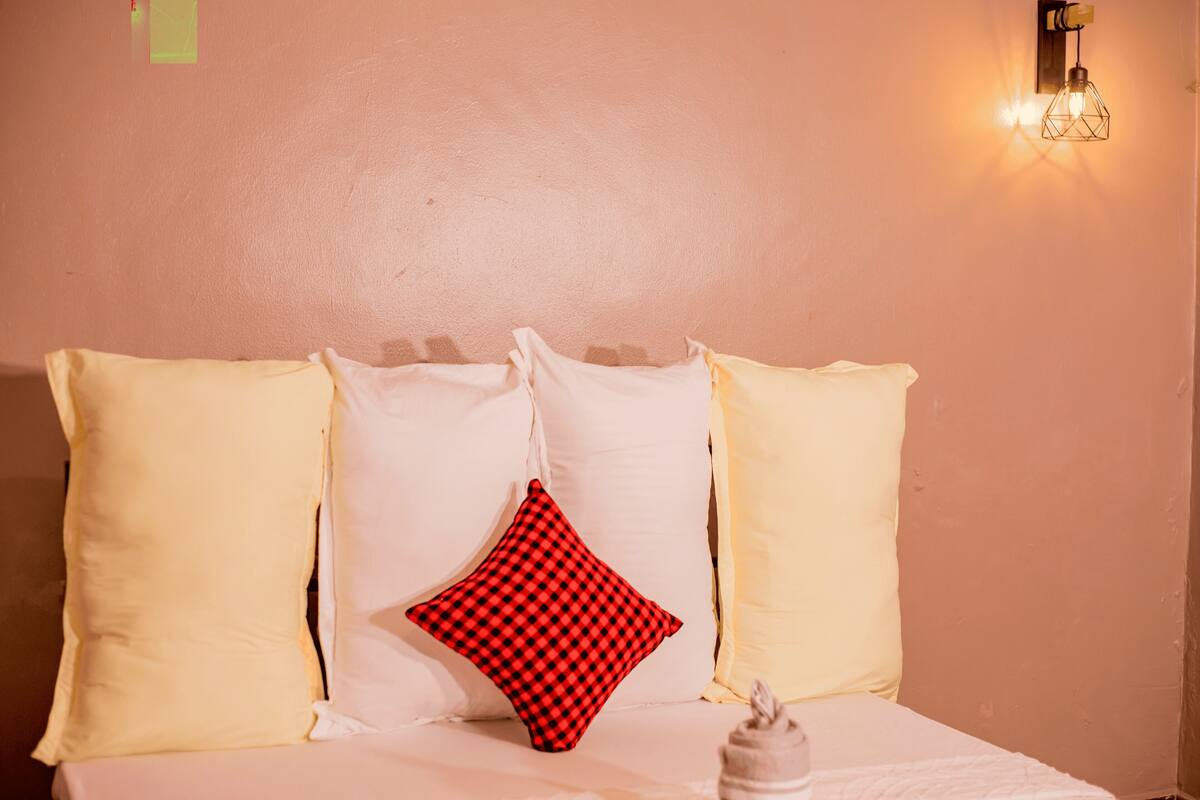 A bed with white and yellow pillows, a red and black checkered pillow, and a wall-mounted light fixture.