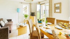 A dining area with wicker chairs, a wooden table set for a meal, a television on a stand, and a window with curtains. - Charming Breton Duplex Cottage (Cancale)
