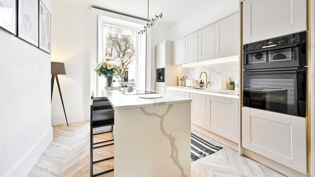 Une cuisine moderne avec des meubles blancs, un îlot en marbre, un four encastré et un luminaire suspendu.