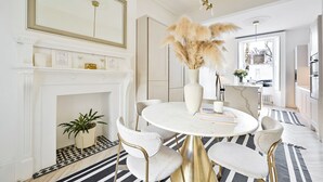 Un espace repas avec une table en marbre blanc, des chaises dorées, un vase contenant de l’herbe des pampas, une cheminée et un grand miroir au-dessus.