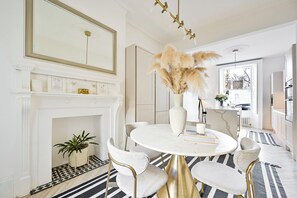A dining area with a white marble table, gold chairs, a vase with pampas grass, a fireplace, and a large mirror above it. - Chelsea Town House, Designer Kitchen + Primary Suite. Minutes to the Kings Road. (London)